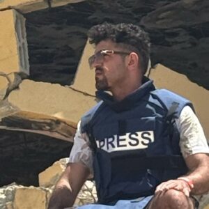 Ido Amiaz sitting in front of a destroyed building wearing a PRESS vest